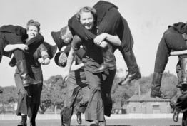 1942 black and white photo from Argus newspaper. Four women of NSW Women's Fire Auxiliary in uniform during drill on grassy oval. Running with dummies resembling fellow members on shoulders. Three women in the foreground smile, enjoying the energetic practice drill.