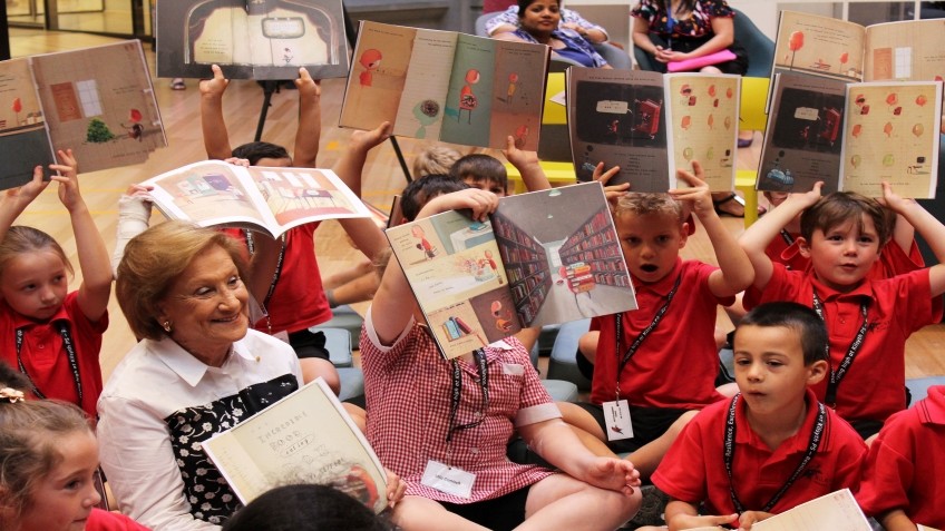 Children in red uniforms hold up books while an older woman smiles at them