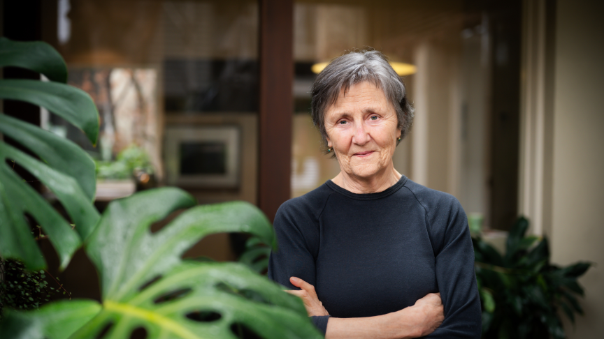 Portrait of author Helen Garner. She is wearing a black long-sleeved top and has her arms crossed. There is a monstera plant in the foreground of the image, on the left side.