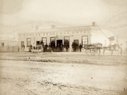 Shows groups of people posed outside Dimmock's Exchange Hotel, with a hitching post and horse trough in the street.